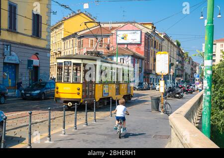 Milan, Italy, September 9, 2018: young boy is riding a bicycle bike near old yellow traditional tram on street with colorful multicolored buildings in beautiful summer day Stock Photo