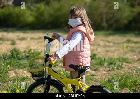 A little girl wearing face mask on a bicycle on a sunny day. Children, play time during the epidemic. Corona virus protection for children. Stock Photo