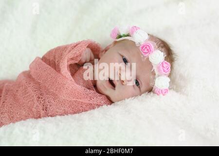 A small newborn girl in a pink diaper and a flower wreath on her head lies on a white blanket and looks at the camera. Tenderness Stock Photo