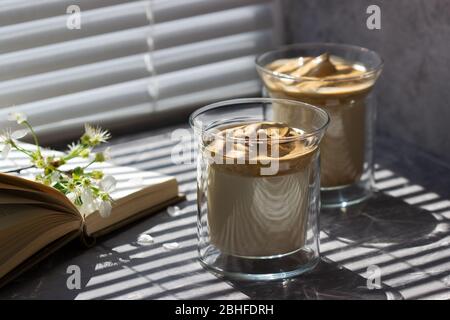 Dalgona Coffee in transparent glasses on a windowsill illuminated by sunlight. Stock Photo