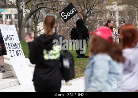 A counter protester dressed as the angel of death waves a sign directed at demonstrators gathered for a 'Rhode Islanders against excessive quarantine' protest in reaction to the state's mandatory stay-at-home order as an attempt to combat COVID-19 at the Rhode Island State House in Providence, Rhode Island on Saturday, April 25, 2020. Rhode Island Governor Gina Raimondo has extended the stay-at-home order in an effort to combat the spread of COVID-19 to May 8th and recently told schools that they will finish the year through via distance learning. Rhode Island has had 6,699 confirmed cases of Stock Photo