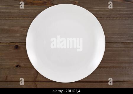 Top down view of a white plate sitting expectantly on a rustic knotted brown wood table top. Stock Photo