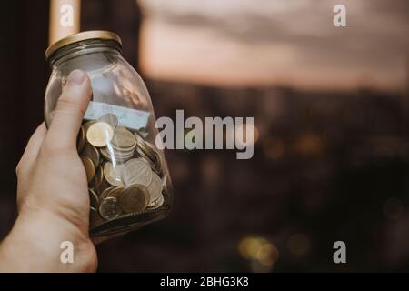 Hispanic man handholding a savings jar with colombian currency bills and coins in a sunset evening Stock Photo