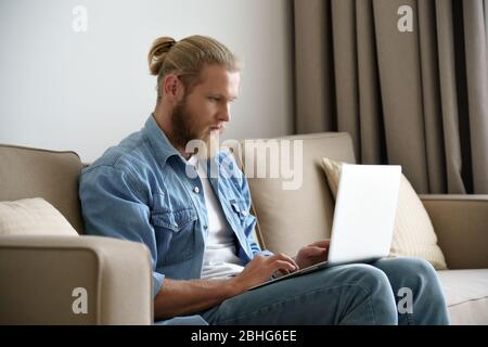 Serious young man working studying from home using laptop sitting on couch. Stock Photo