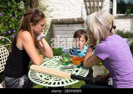 Mother & grandmother outside at table with child boy 3 yrs helping him to eat his lunch food during lockdown in Carmarthenshire Wales UK  KATHY DEWITT Stock Photo