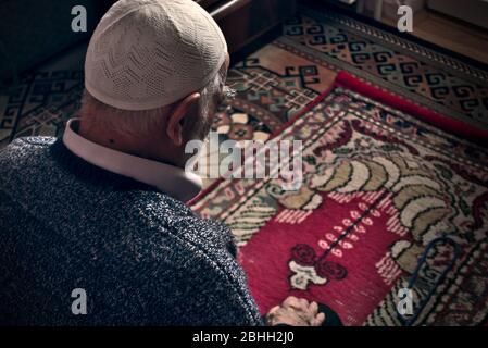 Very old bearded Turkish muslim male at his 80's wearing a prayer cap doing Salah prayer at his home on his prayer rug in Ramadan month Stock Photo