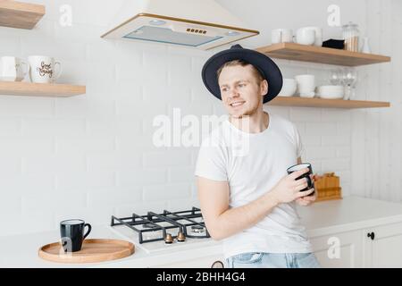 Young man in hat smiles drink fresh coffee in ceramic black mug. Bright kitchen background Stock Photo