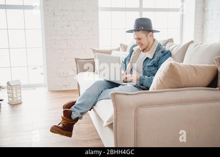 Bearded man coworker hipster in hat working living room home. Male sitting at sofa using laptop and mobile phone. Blurred light background Stock Photo