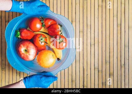 Hands with blue latex gloves holding a blue bowl with different fresh and clean fruits on a wooden base. Bananas, tomatoes, apples, strawberries and o Stock Photo