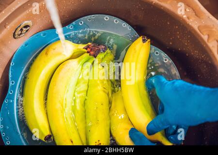 Hands with blue latex gloves disinfecting the bananas in a basin to decontaminate the fruit from the coronavirus. Washing the fruit in the kitchen sin Stock Photo