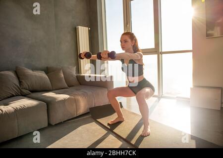 Young girl does gym exercises during sunrise. She is at home due to coronavirus codiv-19 quarantine Stock Photo