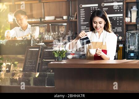Young adult asian female woman barista pouring fresh milk to prepare latte coffee for customer in cafe bar with her colleague working in background. F Stock Photo