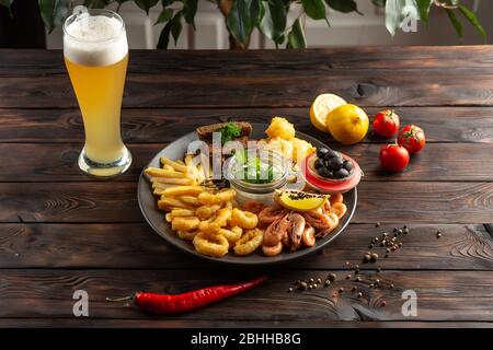 glass of foamy beer and a plate with snacks: shrimp, squid rings, french fries, rye croutons, olives on a wooden background Stock Photo