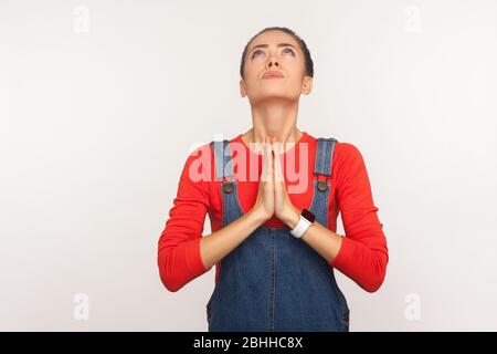 Please god, help! Portrait of imploring worried girl with hair bun in denim overalls looking up and begging, praying with desperate pleading expressio Stock Photo