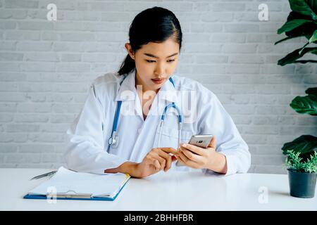 Young Asian female Doctor using a mobile phone at hospital. Healthcare worker scrolling and typing text messages at office. Stock Photo