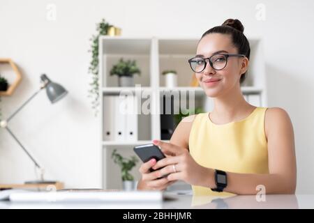 Portrait of content young marketing specialist in yellow dress sitting with smartphone in modern office Stock Photo