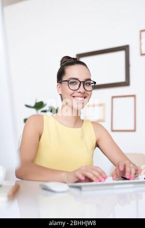 Portrait of smiling beautiful young businesswoman in eyeglasses sitting at desk in modern office and typing on computer Stock Photo