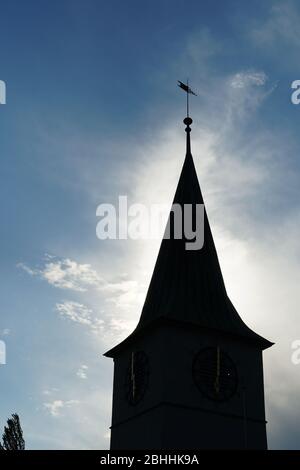 A silhouette of a church tower with a wind pointer on the background of slightly covered sky and surrounded by sunlight Stock Photo