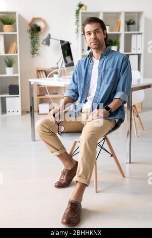 Portrait of content handsome young brunette man in denim shirt sitting on chair and holding eyeglasses in modern office Stock Photo
