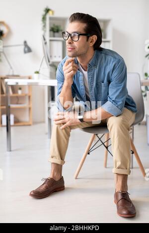 Contemplative young creative manager in eyeglasses sitting on chair in office and leaning head on hand while thinking of new project idea Stock Photo