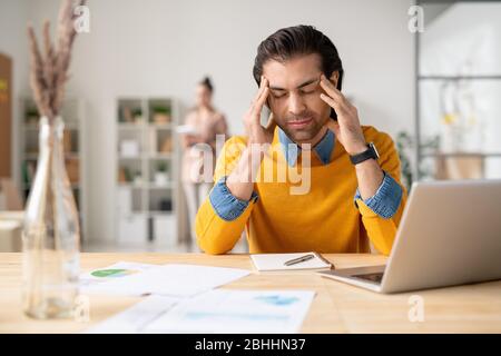 Puzzled young man in yellow sweater sitting at desk with notepad and holding head in hands while suffering from headache at work Stock Photo
