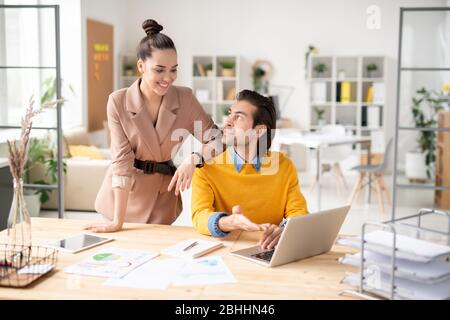 Brunette analyst pointing at laptop screen while discussing statistics with colleague in open space office Stock Photo