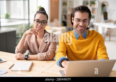 Positive young business colleagues in eyeglasses sitting at table and using laptop while working on web project Stock Photo