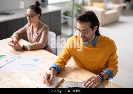 Serious young manager in eyeglasses making notes while analyzing website data on laptop in office Stock Photo