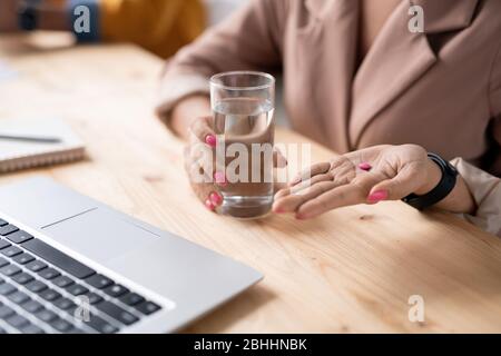 Close-up of unrecognizable businesswoman holding glass of water and taking pink pill at workplace Stock Photo