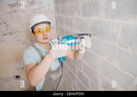Portrait man builder repairer in protective glasses, hard hat, overalls showing having equipment in arm on grey background home brick Stock Photo