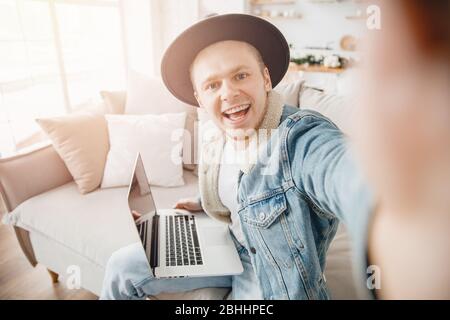 Happy hipster man smile freelancer traveler taking selfie photo in hat, working on laptop home. Concept travel ticket booking Stock Photo