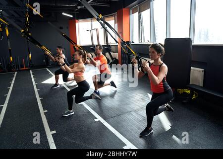 Sporty Lifestyle. Young people doing lunge TRX exercise at gym smiling motivated side view Stock Photo