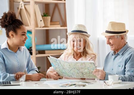 Travel agent at office with senior clients in hats choosing destination on map joyful Stock Photo