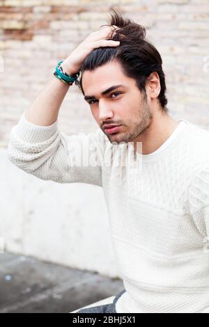 Young handsome man passing his hand through his hair. Trendy hair and stubble beard. Serious attitude. White sweater. Stock Photo