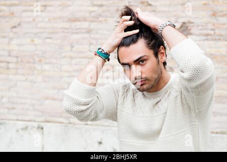 Young handsome man passing his hand through his hair. Trendy hair and stubble beard. Serious attitude. White sweater. Stock Photo