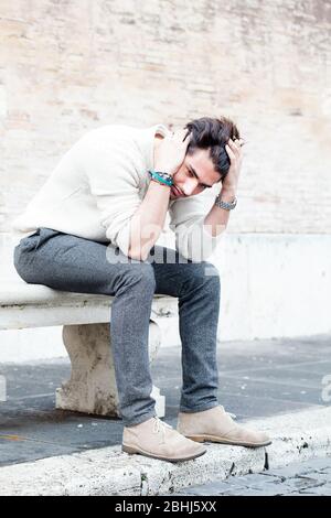 Young worried man sitting and staring outdoors with hands on the head and hair with desperate attitude. White sweater. In the street. Stock Photo