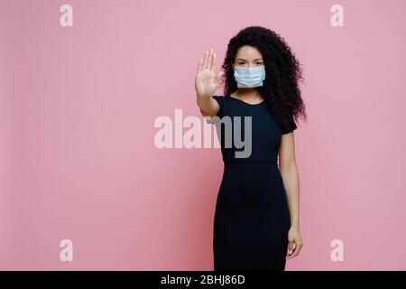 Covid 19 outbreak, viral disease. Photo of ethnic woman with curly hair makes stop gesture with palm, says no to coronavirus, wears protective sterile Stock Photo