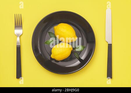 Black plate, knife and fork with lemon on yellow background. Top view. Stock Photo