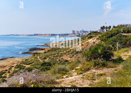 Dehesa de Campoamor high rise residential building town, view from Cabo Roig beach rocky seaside. Calm blue Mediterranean Sea, sunny day, no people. Stock Photo