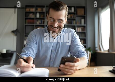 Smiling businessman wearing glasses using phone, writing in notebook Stock Photo