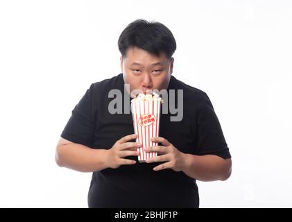 Funny young asian fat man in black polo shirt eating popcorn isolated over white background. Stock Photo