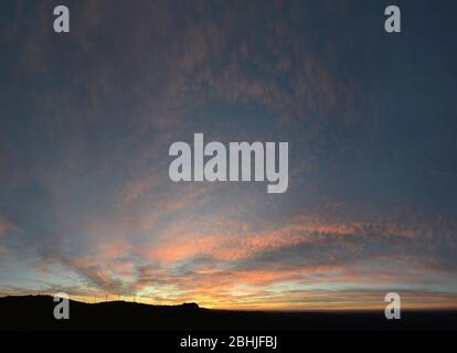 The Vast Amplitude of the Sunset Sky dotted with little orange clouds and the silhouette of distant hills and wind turbines Stock Photo