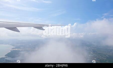 wing of airplane flying through clouds over receding land with towns rivers mountains against sky with white clouds Stock Photo