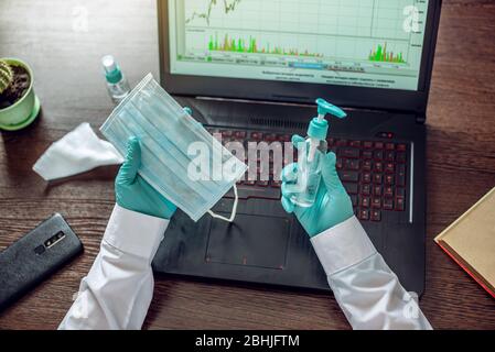 A woman in rubber gloves holding a medical mask and a sanitizer at her Desk over her laptop in the office. Cleanliness and hygiene in the fight agains Stock Photo