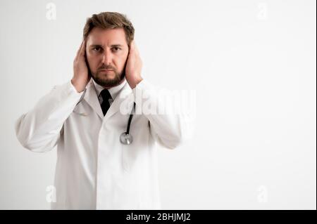 Portrait of male doctor with stethoscope in medical uniform covering ears like deaf concept posing on a white isolated background Stock Photo