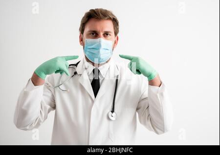 Portrait of male doctor with stethoscope in medical uniform wears a protective mask, with his fingers pointed posing on a white isolated background Stock Photo