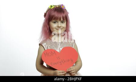 Smiling little girl princess holds a cut red paper heart with text message about mom in her hands. White background. Cute child enjoying happy childhood, showing love sign. Mother's day concept Stock Photo