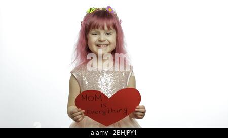 Smiling little girl princess holds a cut red paper heart with text message about mom in her hands. White background. Cute child enjoying happy childhood, showing love sign. Mother's day concept Stock Photo