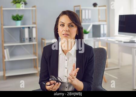 Serious businesswoman in a black suit looking at the camera talking to a partner while sitting in the office. Stock Photo