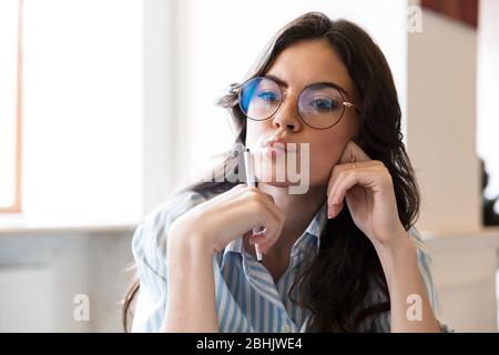 Attractive pensive young brunette woman studying in the cafe indoors, holding pen at her face Stock Photo